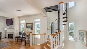 A spacious living room with hardwood flooring, a fireplace, a dining area with chairs, and a staircase with a black railing. - 22 Coast Cottages by Hodnett Cooper (St. Simons Island)