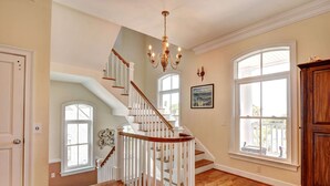 A spacious room with hardwood flooring, a white staircase with wooden railings, and large windows offering views of the outdoors. - 2225 Bruce Drive by Hodnett Cooper (St. Simons Island)
