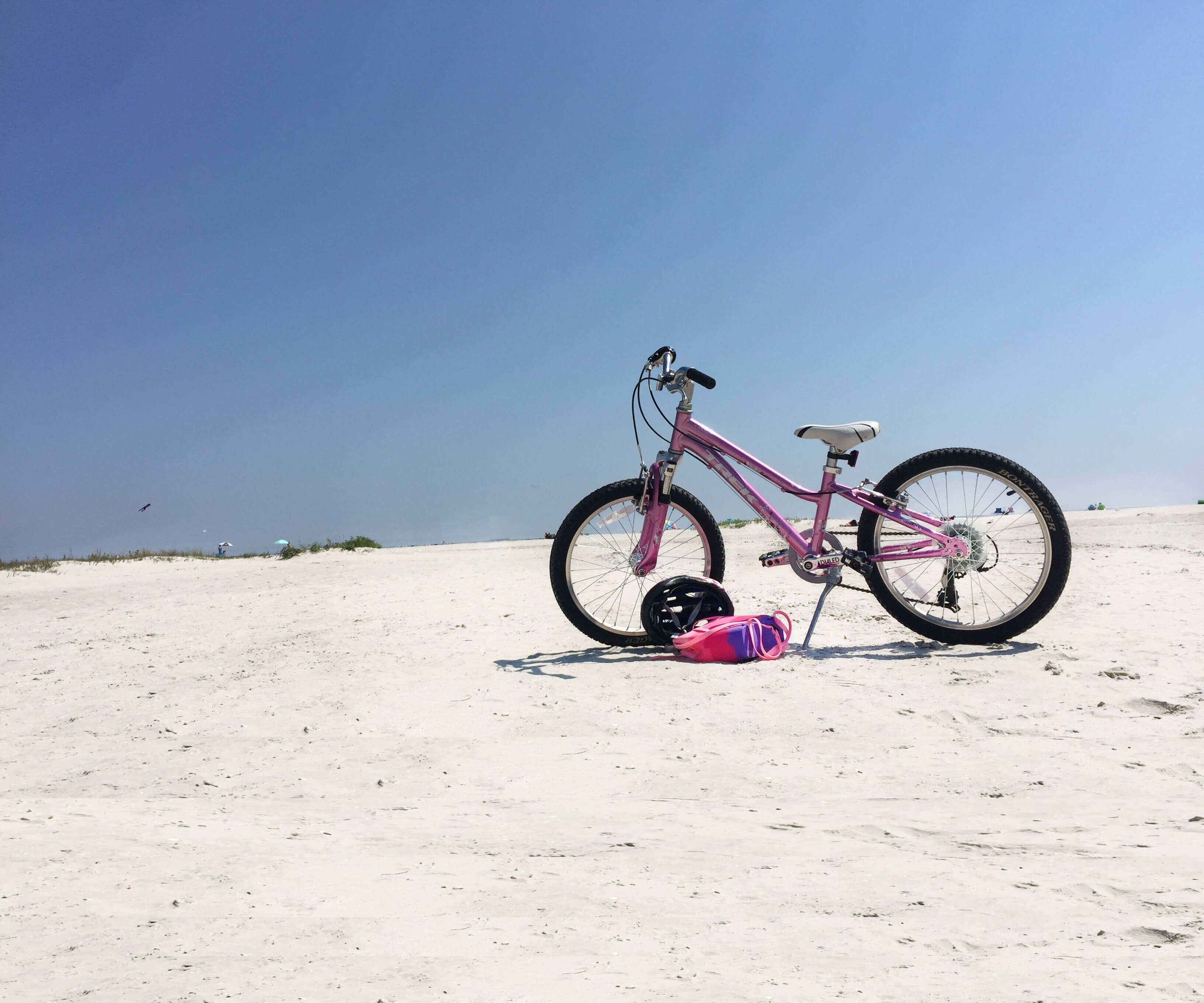 Ein pinkfarbenes Fahrrad mit Helm und Tasche auf einem Sandstrand.