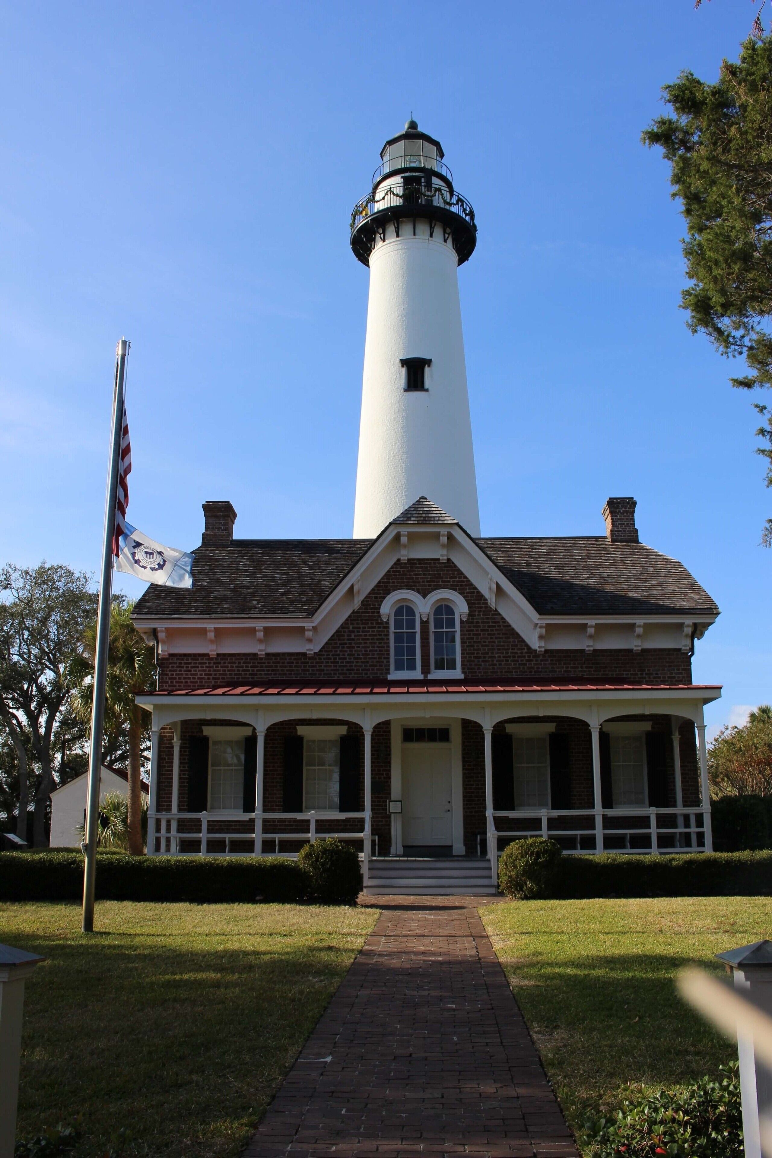 A lighthouse with a brick base and a white tower, a flagpole with an American flag, and a well-maintained lawn.