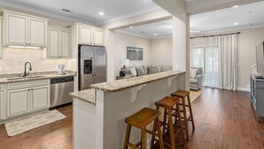 A modern kitchen with white cabinets, a granite countertop, and a stainless steel refrigerator. There are wooden stools at the island and a rug on the floor. - 1203 Reserve Lane by Hodnett Cooper (St. Simons Island)