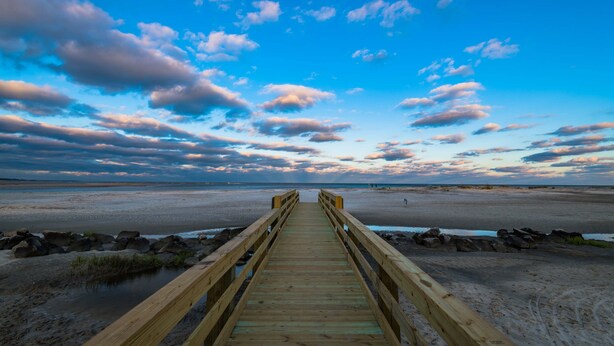 A wooden boardwalk leading to a beach with a clear sky and scattered clouds.