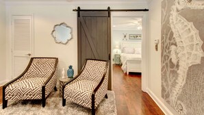 A room with two patterned armchairs, a white door, a mirror, and a sliding barn door leading to another room with a bed and a decorative wall.