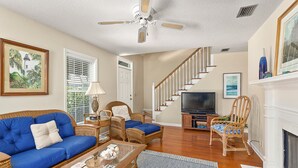 A living room with a blue sofa, wicker chairs, a television, and a fireplace. - 111 Mews Circle by Hodnett Cooper (St. Simons Island)
