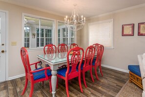 A dining room with a glass table surrounded by six red chairs with blue cushions, a white ceiling fan, and a chandelier. - 4316 7th Street by Hodnett Cooper (St. Simons Island)