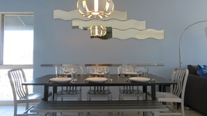 Un comedor con una mesa rectangular dispuesta para cuatro personas, rodeada de sillas blancas. Un candelabro moderno cuelga sobre la mesa, y un espejo grande está montado en la pared encima.