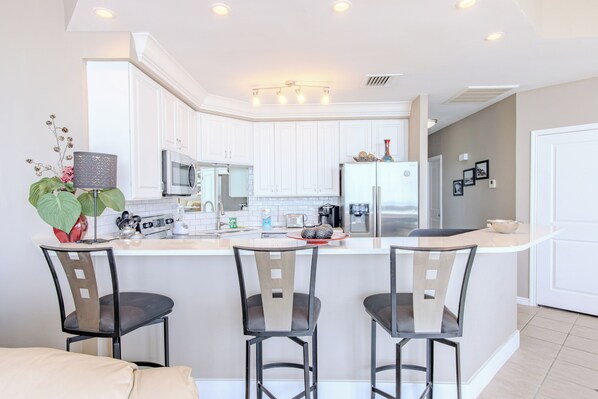 Una cocina moderna con armarios blancos, una isla central con taburetes y electrodomésticos de acero inoxidable, además de una planta decorativa.