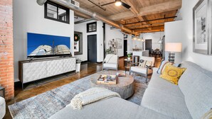 A modern living room with a large sofa, a coffee table, and a TV on a white cabinet. There is a ceiling fan, a window, and a brick wall. - Urban Oasis In Downtown Waco; short walk to Silos (Waco)