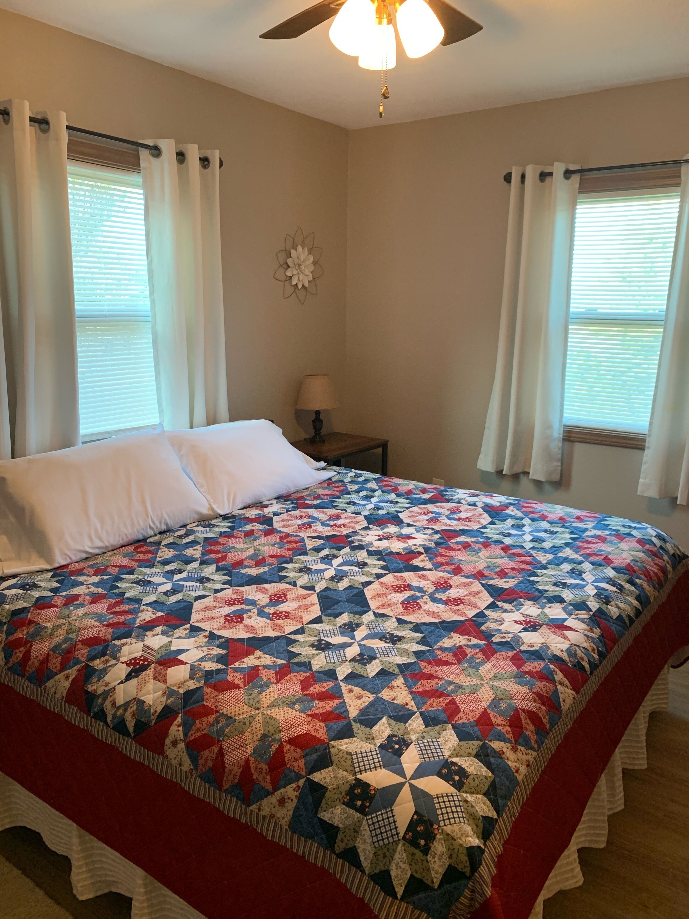 A bedroom with a large bed, white curtains, a ceiling fan, and a window with blinds.