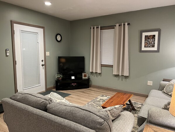 Un salon avec un canapé gris, une table basse, un téléviseur, une fenêtre avec des rideaux, une horloge et un tableau encadré au mur.