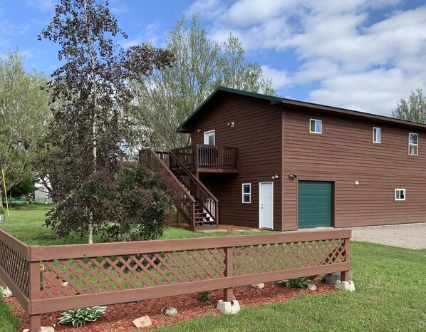 Un immeuble brun à deux étages, avec une porte de garage verte, un escalier en bois menant à un balcon et une clôture en bois au premier plan.