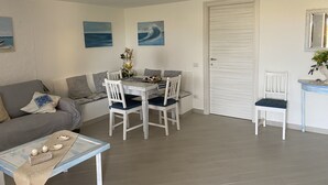 Una sala de estar con una mesa de comedor, sillas, un sofá y una mesa de centro. Hay cuadros en la pared y una puerta al fondo.
