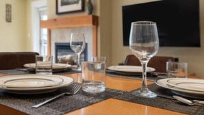 Una mesa de comedor dispuesta con platos, vasos y cubiertos, con una chimenea y un cuadro enmarcado al fondo.