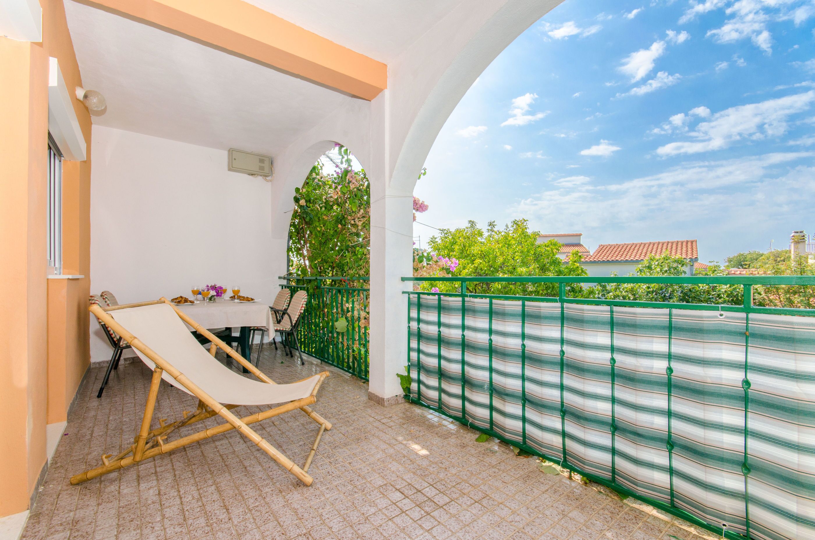 Une terrasse couverte avec une balançoire en bambou, une table avec des chaises et une vue sur une clôture verte et des arbres.