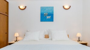 A hotel room with a bed, two bedside lamps, a painting of a boat on the wall, and a wooden wardrobe. - Apartments Zoric (Rogoznica)