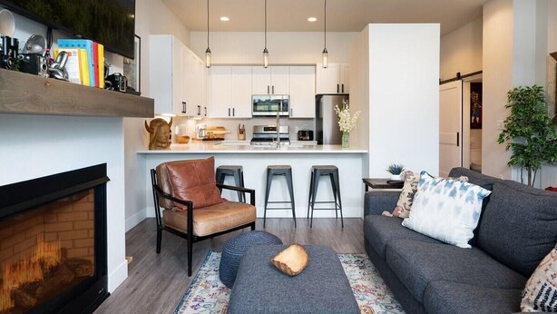 A modern living room with a fireplace, sofa, ottoman, and a kitchen area with bar stools. - Hideaway Station TS 23 - Downtown Winter Park (Winter Park)