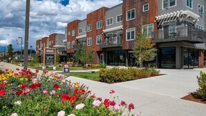 A modern multi-story building with a mix of brick and metal exterior, a landscaped courtyard with colorful flowers, and a sign indicating a nearby hotel. - Hideaway Station TS 23 - Downtown Winter Park (Winter Park)