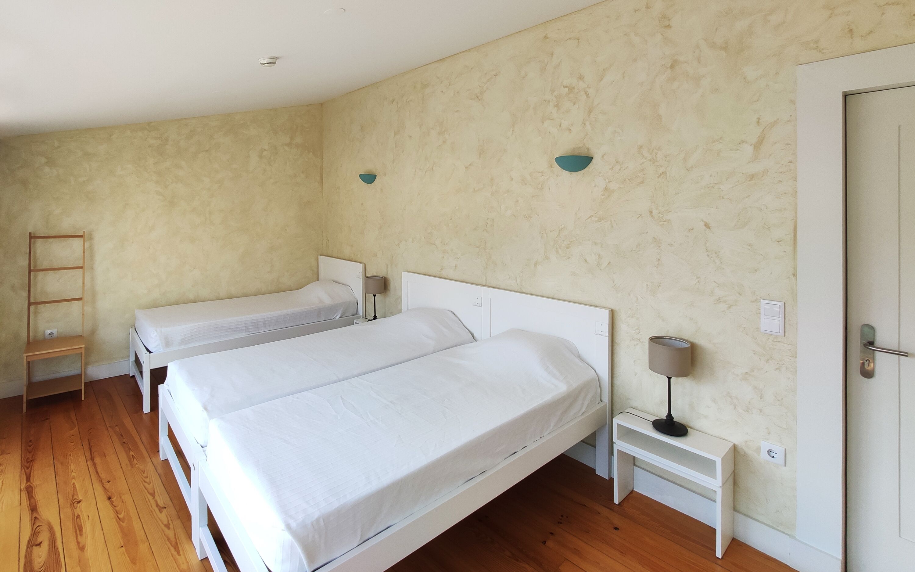 Een kamer met twee eenpersoonsbedden, een houten stoel, een nachtkastje met een lamp en een deur.