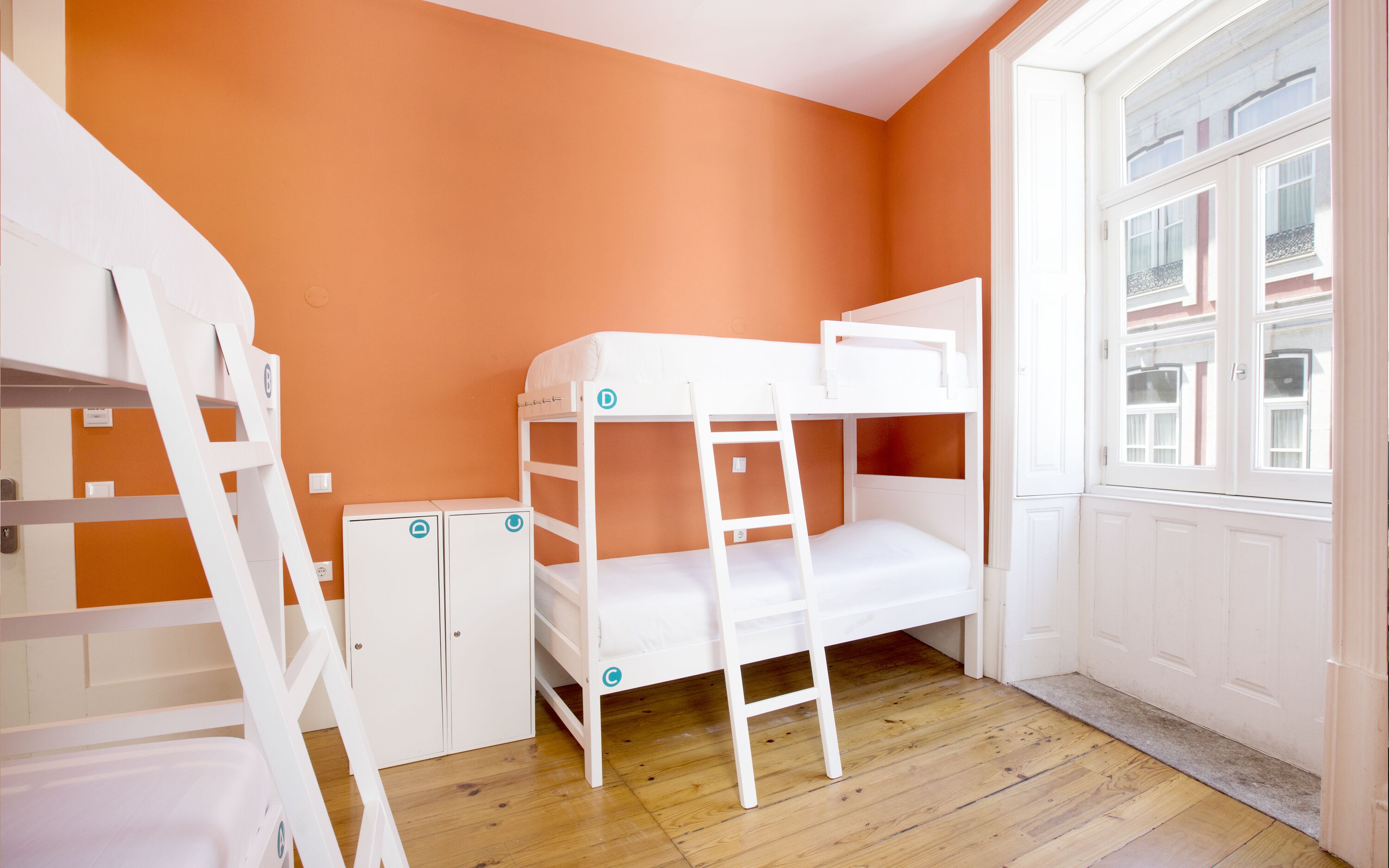 A bunk bed room with orange walls, a wooden floor, and a window with a view of buildings.
