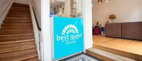 A reception area with a 'Best Guest Hostel' sign, wooden stairs, a patterned rug, and a sofa in the background.