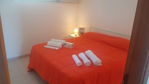 Una cama bien tendida con una colcha naranja, toallas blancas y una mesita de noche con lámpara.
