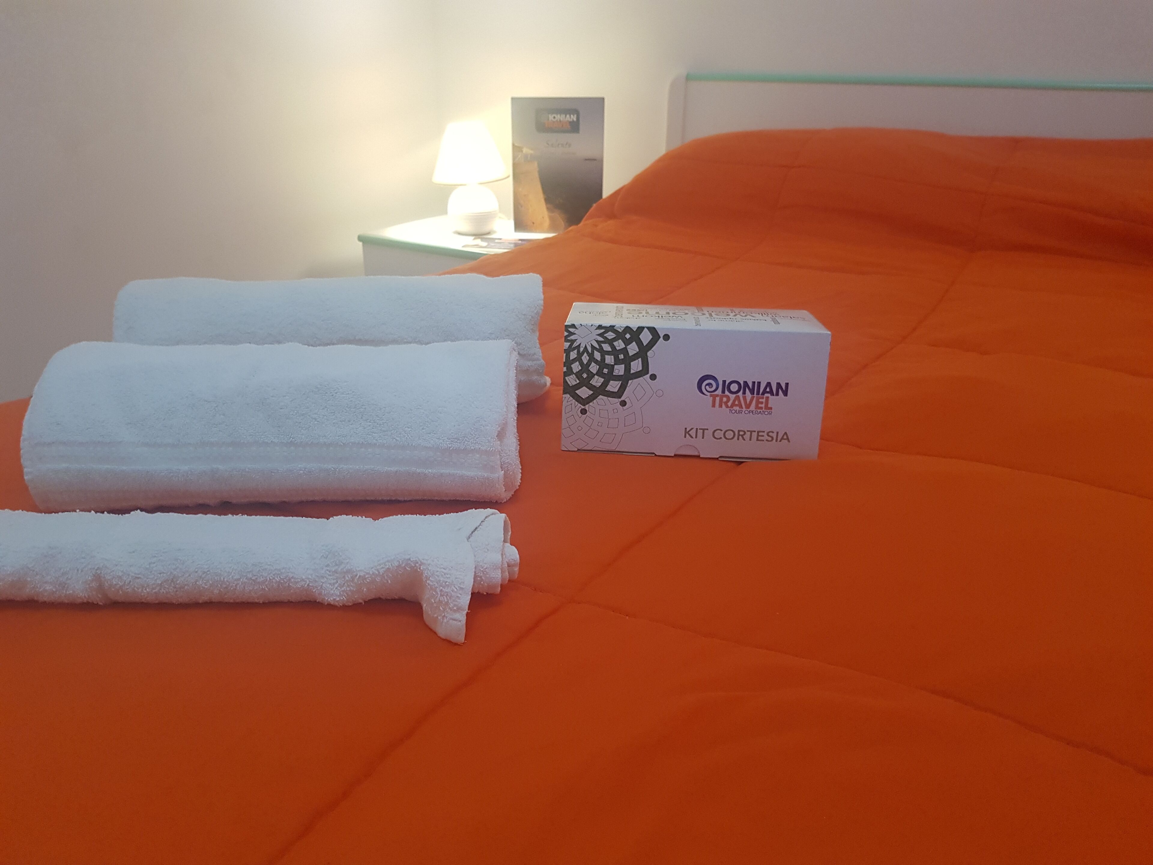 En seng med et orange lagen, to pænt foldede hvide håndklæder og en æske med teksten "IONIAN TRAVEL TOUR OPERATOR KIT CORTEZIA" på et natbord.