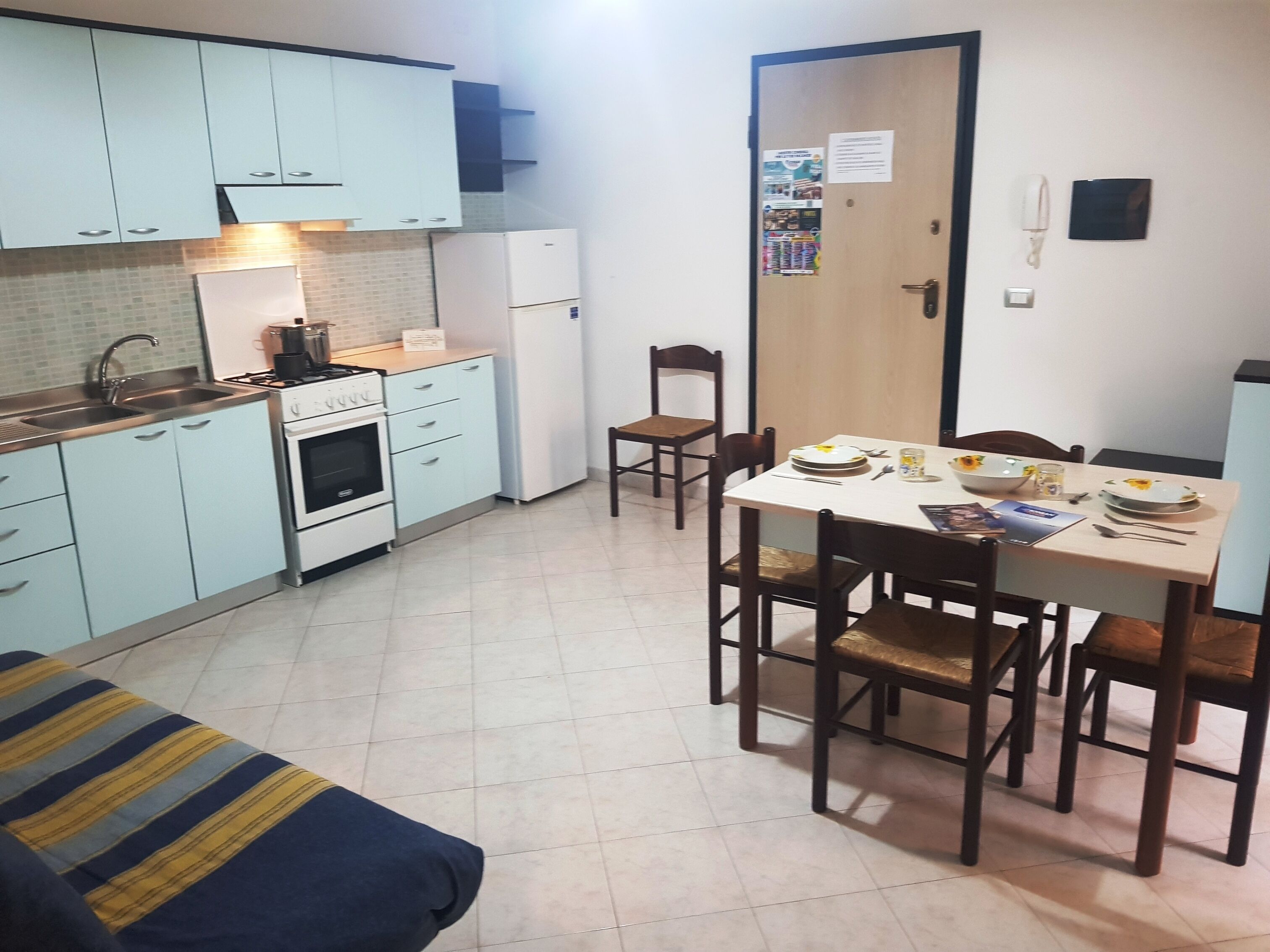 A compact kitchen with light blue cabinets, a stove, and a refrigerator. There is a dining table with chairs and a small sofa in the foreground.