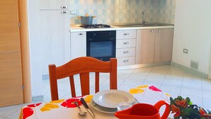 Cocina compacta con gabinetes blancos, estufa y lavamanos. En primer plano, una mesa de comedor con mantel floral y una jarra roja.