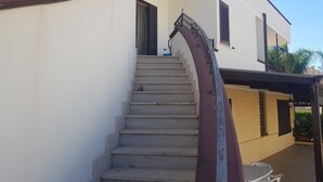 Una escalera que lleva a la entrada de un edificio, con una zona exterior cubierta y una pequeña mesa y sillas cerca.