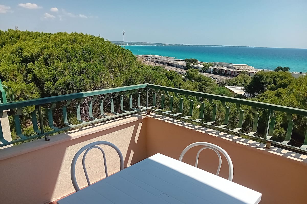 A balcony with a table and chairs, overlooking a coastal area with buildings and trees.