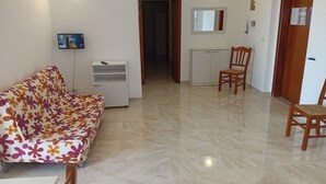 Una sala de estar con un sofá, un pequeño armario blanco, una silla de madera y un televisor en la pared.