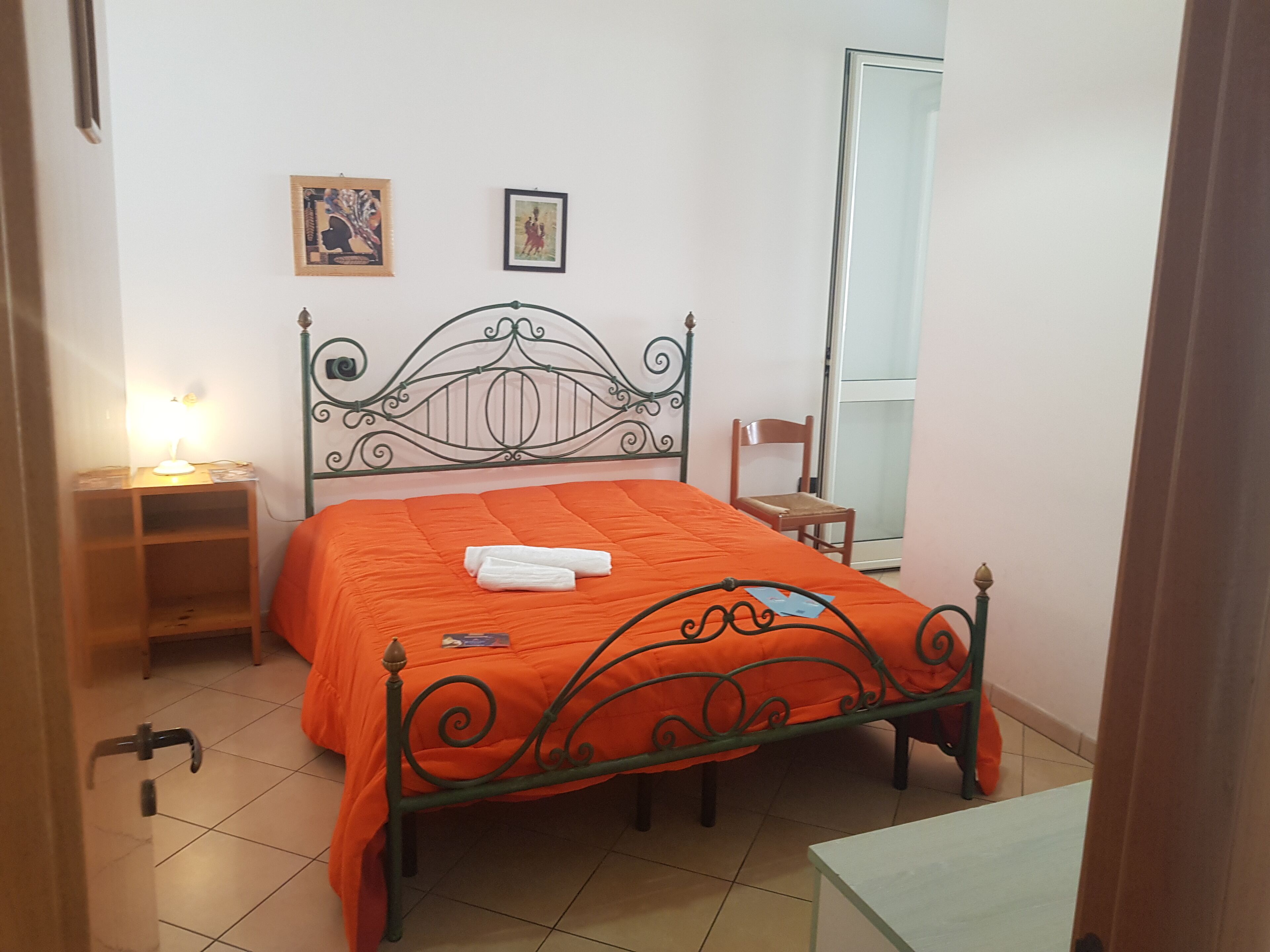Un letto rifatto con una coperta arancione, un comodino con una lampada e una sedia appoggiata al muro.