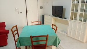 Un área de comedor con una mesa cubierta con mantel verde, sillas de madera, un sofá rojo, un televisor sobre un mueble bajo y un aparador de vidrio con vajilla.