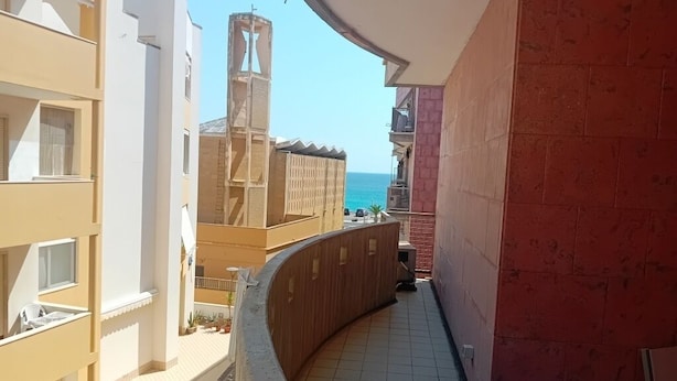 Un pasillo con piso de baldosas, barandilla curva y vista al mar y edificios en la distancia.