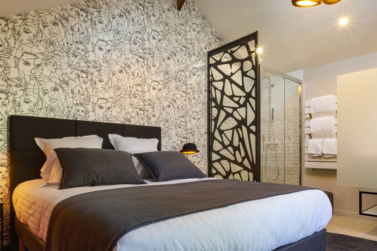 Une chambre d’hôtel moderne dotée d’un grand lit, d’une tête de lit décorative, d’une douche en verre et d’un mur orné d’un motif original.