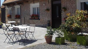 Un bâtiment en briques avec un toit en tuiles, une porte noire et des fenêtres ornées de jardinières. Il y a des places assises à l'extérieur, avec une table et des chaises.