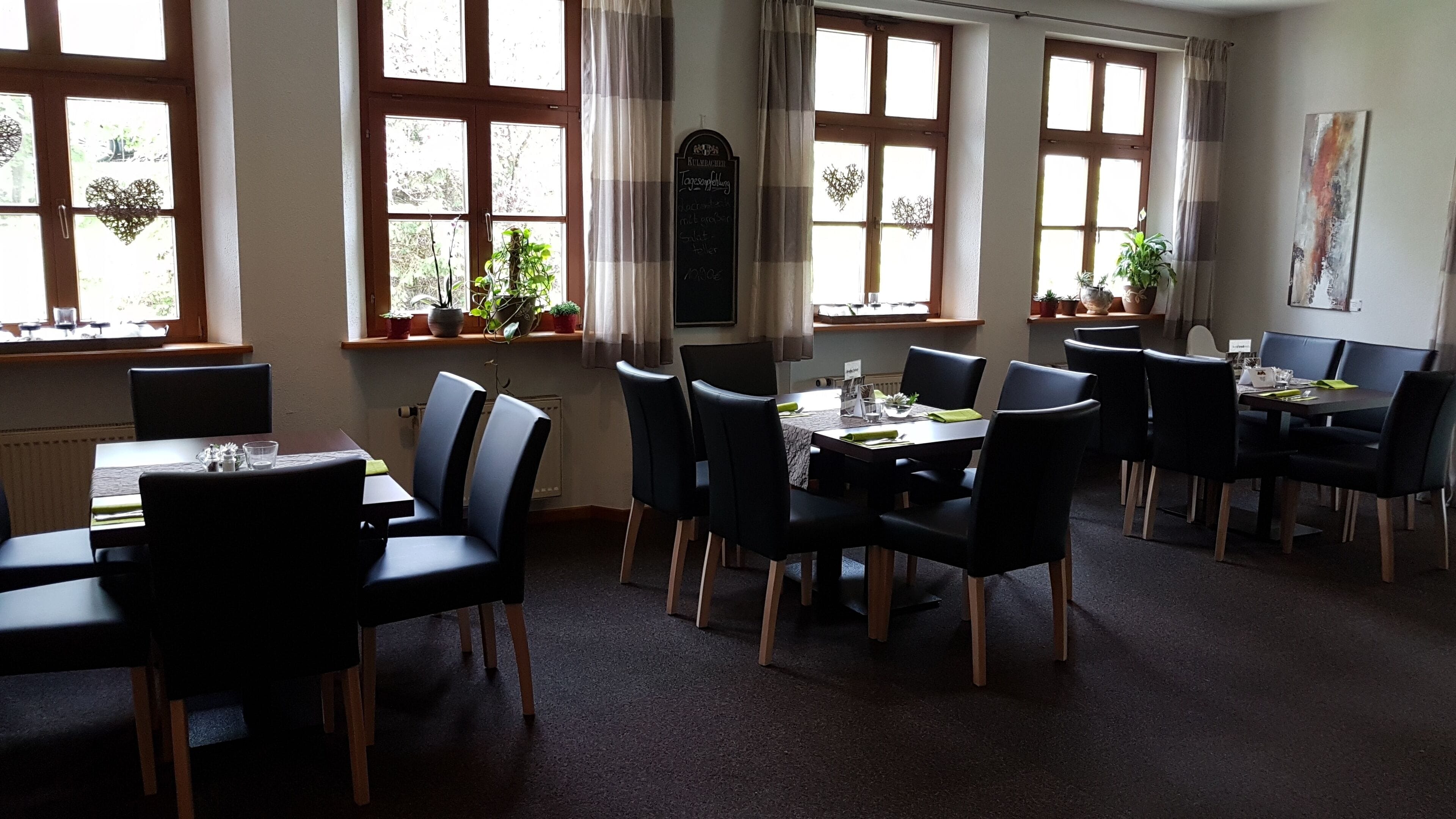 A dining area with multiple tables and chairs, large windows with curtains, and potted plants on the windowsills.