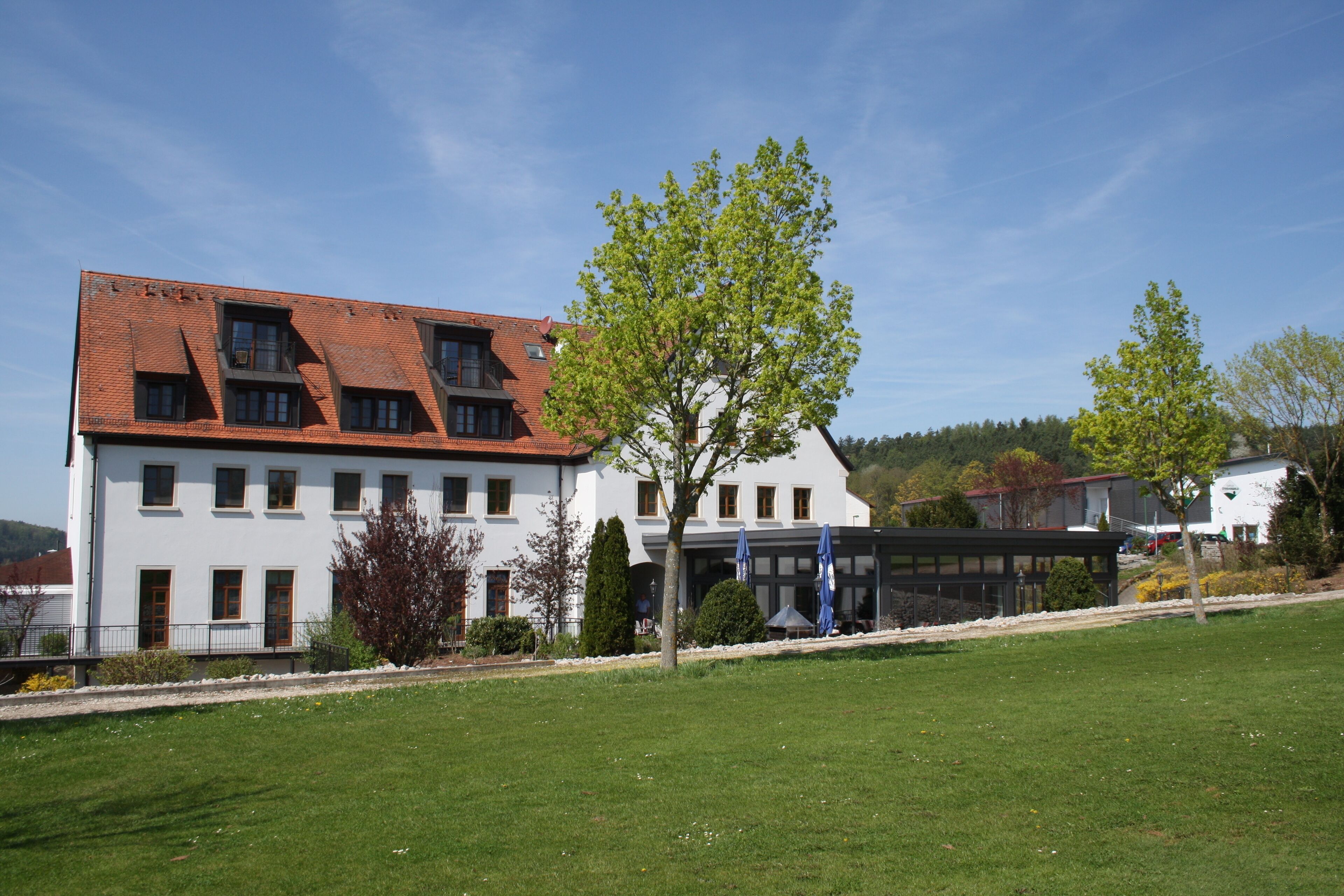 A large, white building with a red-tiled roof, multiple windows, and a covered outdoor area with umbrellas.
