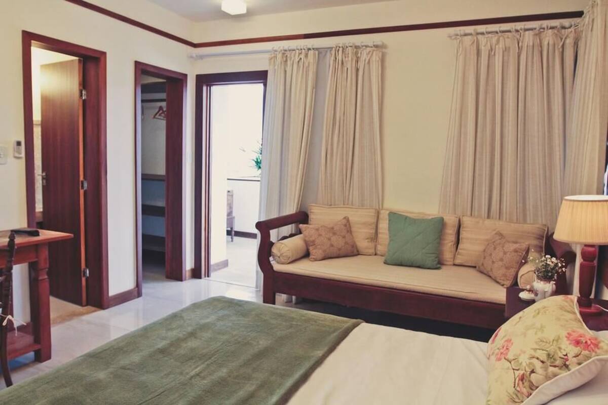 A room with a bed, a sofa, a table, and a lamp. There are curtains on the windows and a door leading to another room.