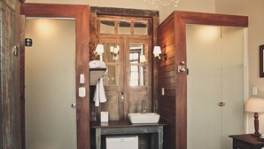 Habitación de hotel pequeña con puerta de madera, un lavamanos, un espejo, una cama y una mesita de noche con lámpara.
