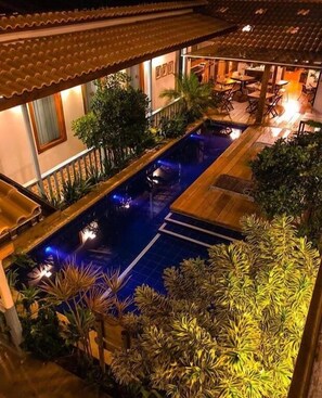 A rooftop pool area with a wooden deck, surrounded by lush greenery and a tiled roof.