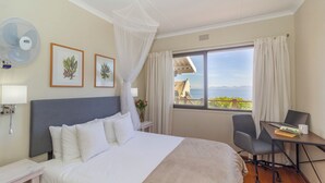 Une chambre d’hôtel avec un lit, un bureau avec un ordinateur portable, une chaise, un ventilateur et une fenêtre donnant sur la mer.