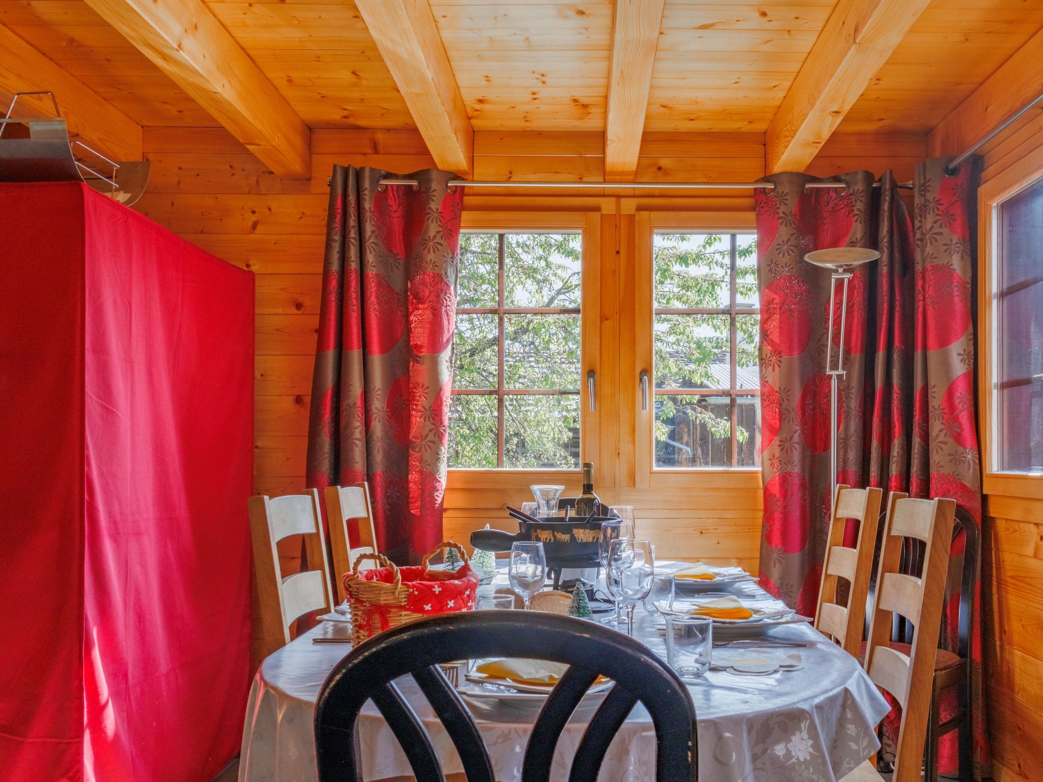 Une salle à manger aménagée dans une cabane en bois, avec une table dressée pour quatre personnes, une fenêtre avec des rideaux et une vue sur les arbres à l’extérieur.
