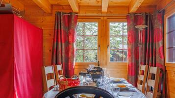 Une salle à manger aménagée dans une cabane en bois, avec une table dressée pour quatre personnes, une fenêtre avec des rideaux et une vue sur les arbres à l’extérieur.