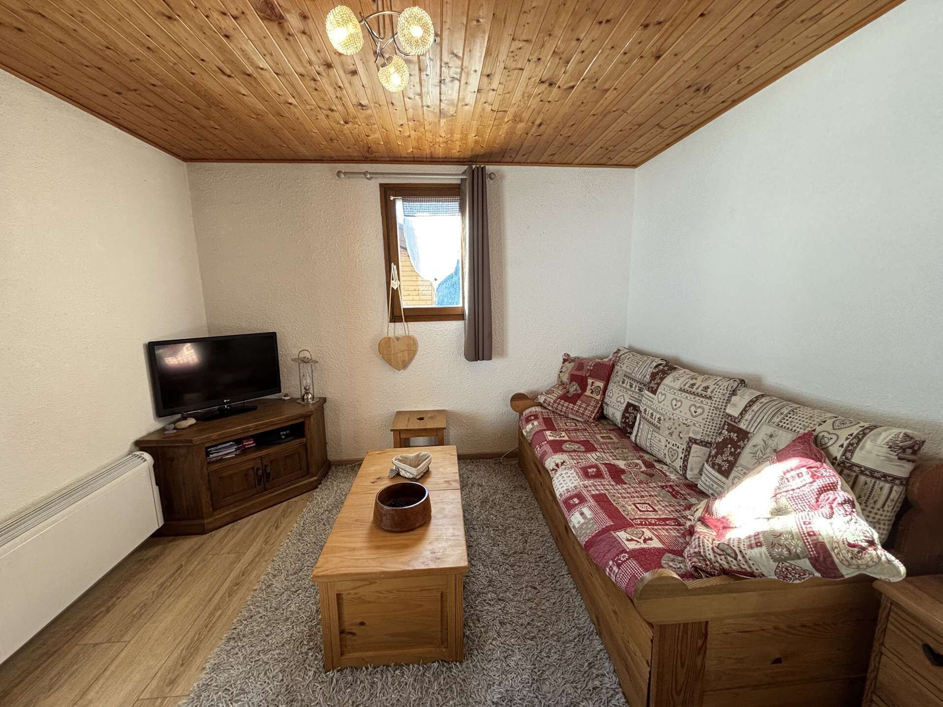 Un salon chaleureux avec un plafond en bois, un téléviseur posé sur un meuble en bois, un canapé agrémenté de coussins à motifs et une petite table sur laquelle se trouve un bol.