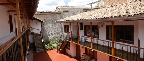 A courtyard with a wooden staircase, a covered seating area with chairs, and a stone wall in the background.