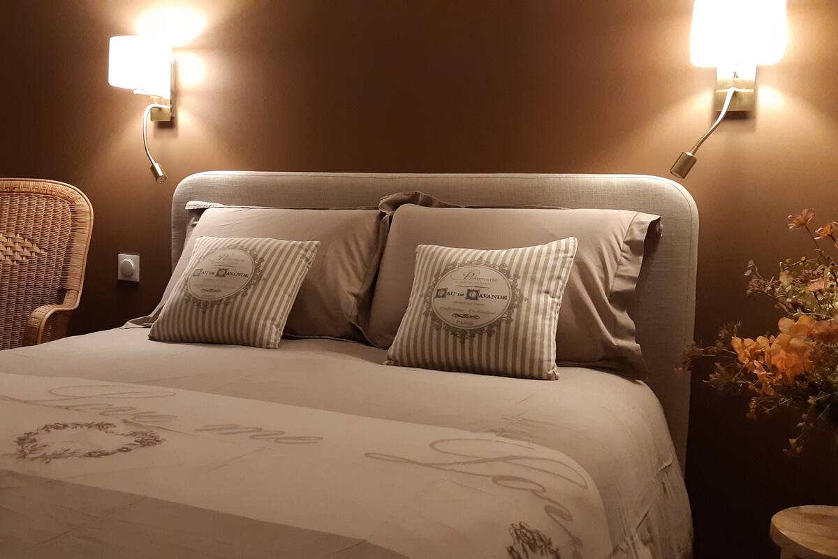 A neatly made bed with a floral bedspread, two pillows with a face design, a wicker chair, and a wall-mounted lamp.