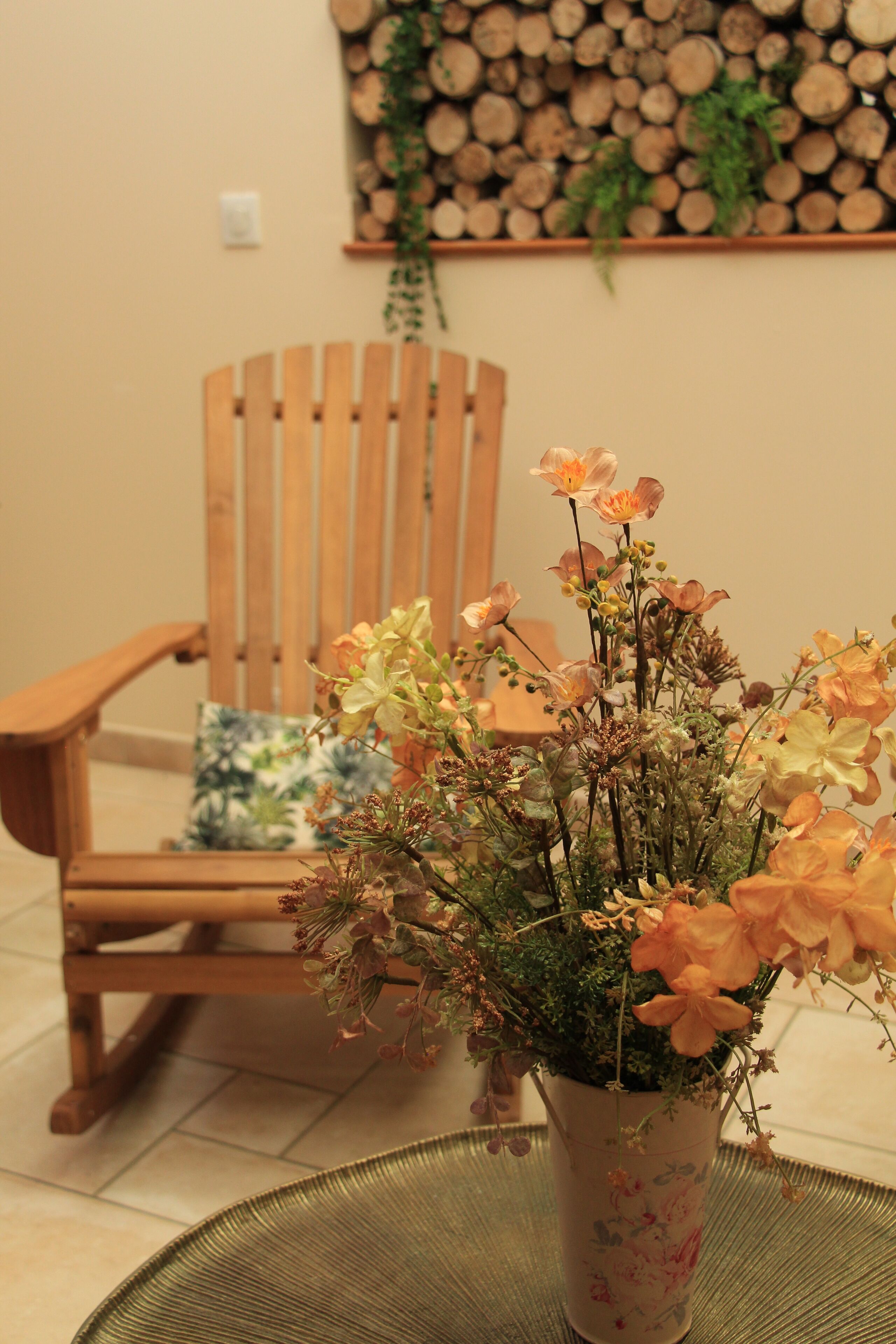 Une chaise en bois avec un coussin, un arrangement floral dans un pot décoré, et un mur orné de bûches empilées et de verdure.