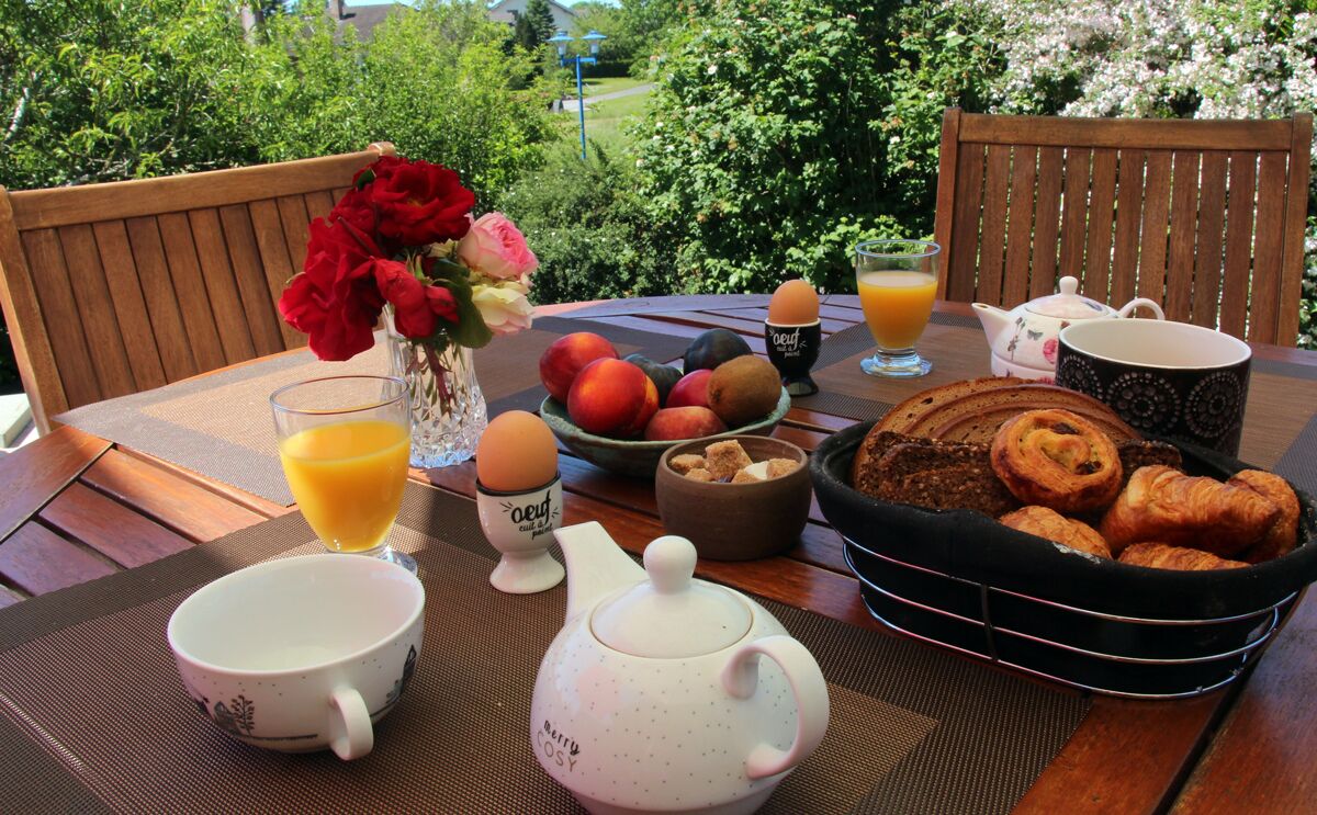 A breakfast setup with a teapot, cups, a fruit bowl, a basket of croissants, and a vase of flowers on an outdoor table.