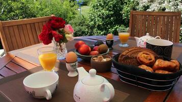 Een ontbijtarrangement met een theepot, kopjes, een schaal fruit, een mandje croissantjes en een bloevas op een buitentafel.